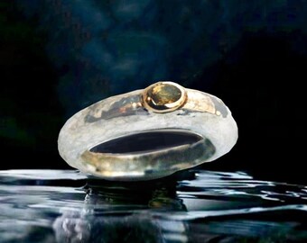 May include: A silver ring with a gold band and a small, round, yellow gemstone. The ring is set against a dark, blurred background, with reflections in the water.
