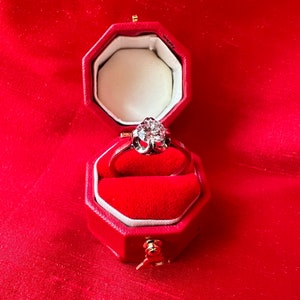 May include: A diamond ring in a red octagonal jewelry box. The ring features a large, sparkling diamond set in a silver band. The box is lined with red velvet and sits against a red background.