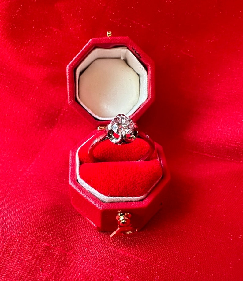 May include: A diamond ring in a red octagonal jewelry box. The ring features a large, sparkling diamond set in a silver band. The box is lined with red velvet and sits against a red background.