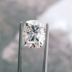 May include: A brilliant, cushion-cut diamond held by metal tweezers. The diamond's facets reflect light, creating a sparkling effect. The gemstone is set against a blurred, neutral background, highlighting its clarity and brilliance. A close-up view of a precious stone.