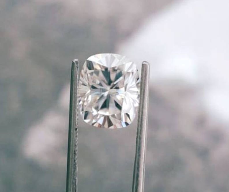 May include: A brilliant, cushion-cut diamond held by metal tweezers. The diamond's facets reflect light, creating a sparkling effect. The gemstone is set against a blurred, neutral background, highlighting its clarity and brilliance. A close-up view of a precious stone.