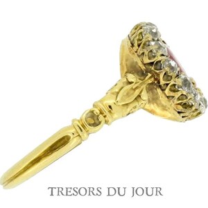 Peut inclure: Une bague en or avec une pierre centrale rouge, entour&eacute;e de petites pierres claires. La bague pr&eacute;sente un design orn&eacute; et une longue et fine monture. Le texte "TRESORS DU JOUR" est visible en bas de l'image.