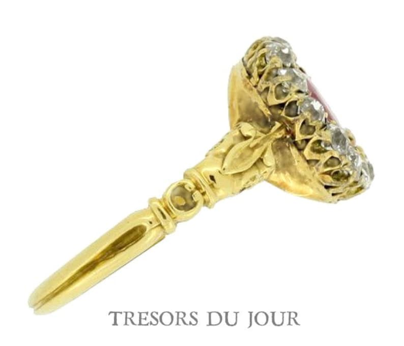 Peut inclure: Une bague en or avec une pierre centrale rouge, entour&eacute;e de petites pierres claires. La bague pr&eacute;sente un design orn&eacute; et une longue et fine monture. Le texte "TRESORS DU JOUR" est visible en bas de l'image.