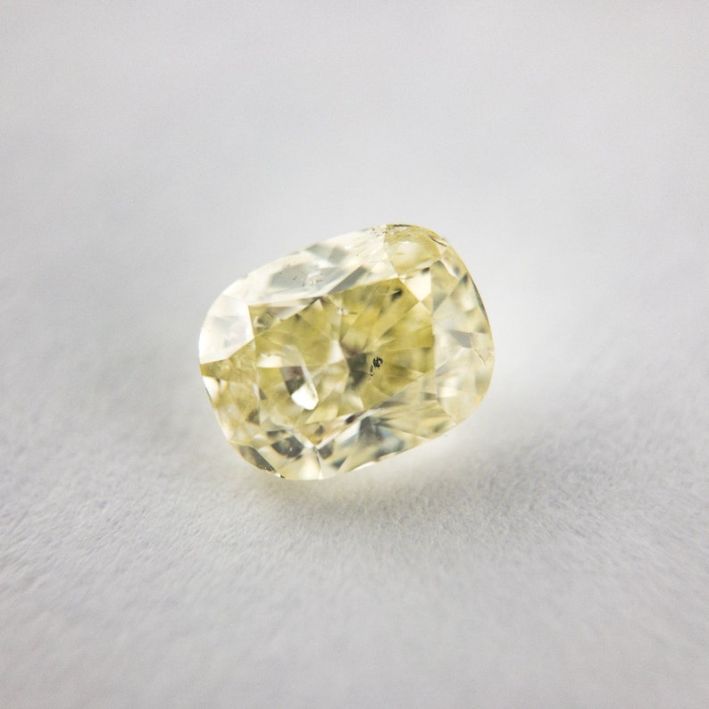 May include: A pale yellow, faceted gemstone with a cushion cut. The gemstone is set against a white background.