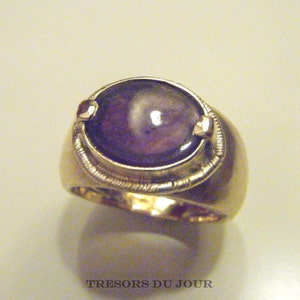 Op de afbeelding: Een gouden ring met een grote, ovale, paarse edelsteen die in een bezel-setting is gezet. De ring heeft een getextureerde band en twee kleine gouden accenten aan weerszijden van de edelsteen.