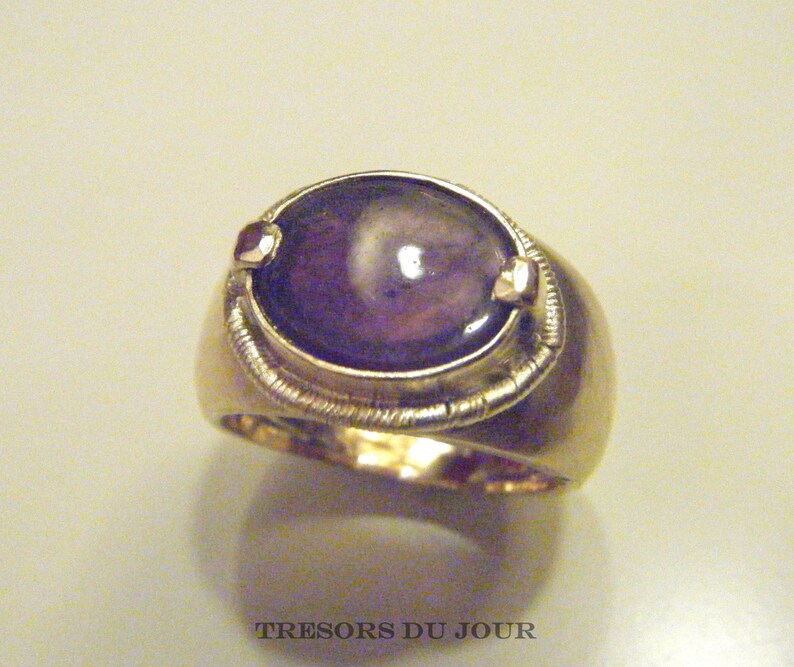 Op de afbeelding: Een gouden ring met een grote, ovale, paarse edelsteen die in een bezel-setting is gezet. De ring heeft een getextureerde band en twee kleine gouden accenten aan weerszijden van de edelsteen.