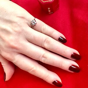 May include: A silver ring with a large, round, clear gemstone and smaller surrounding stones. The ring is on a hand with dark red nail polish, set against a red background. A red jewelry box with a gold trim is also visible.