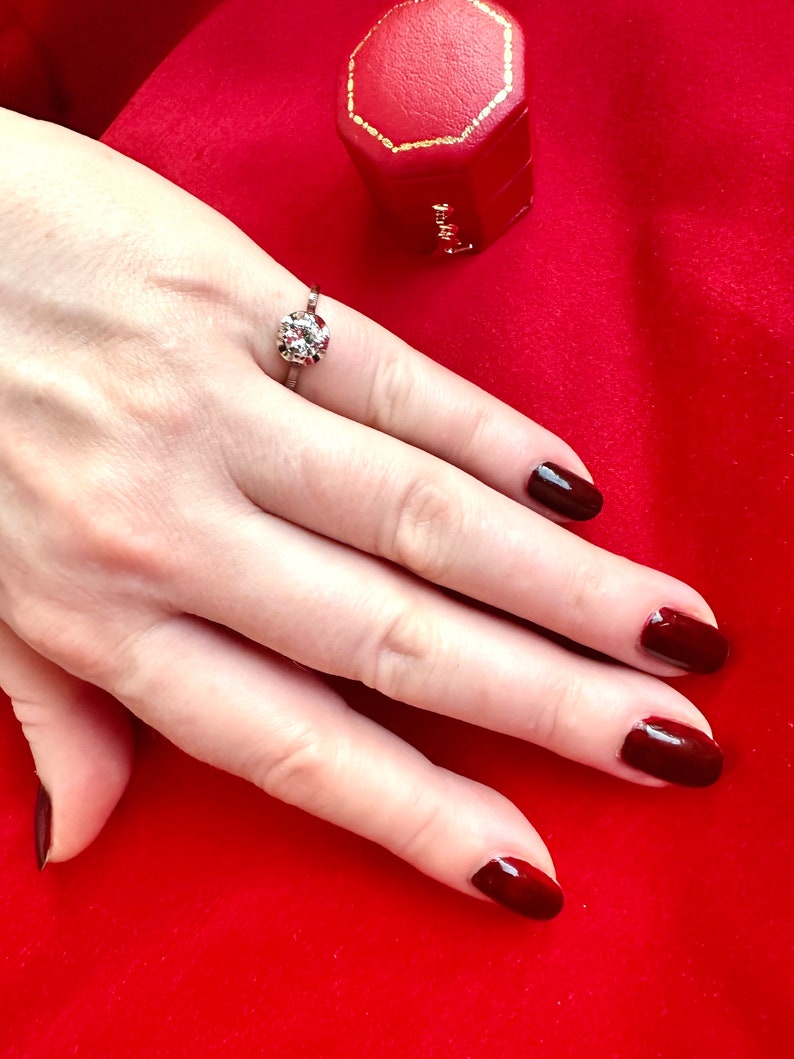 May include: A silver ring with a large, round, clear gemstone and smaller surrounding stones. The ring is on a hand with dark red nail polish, set against a red background. A red jewelry box with a gold trim is also visible.