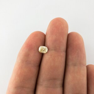 May include: A light yellow cushion-cut gemstone held between two fingers.