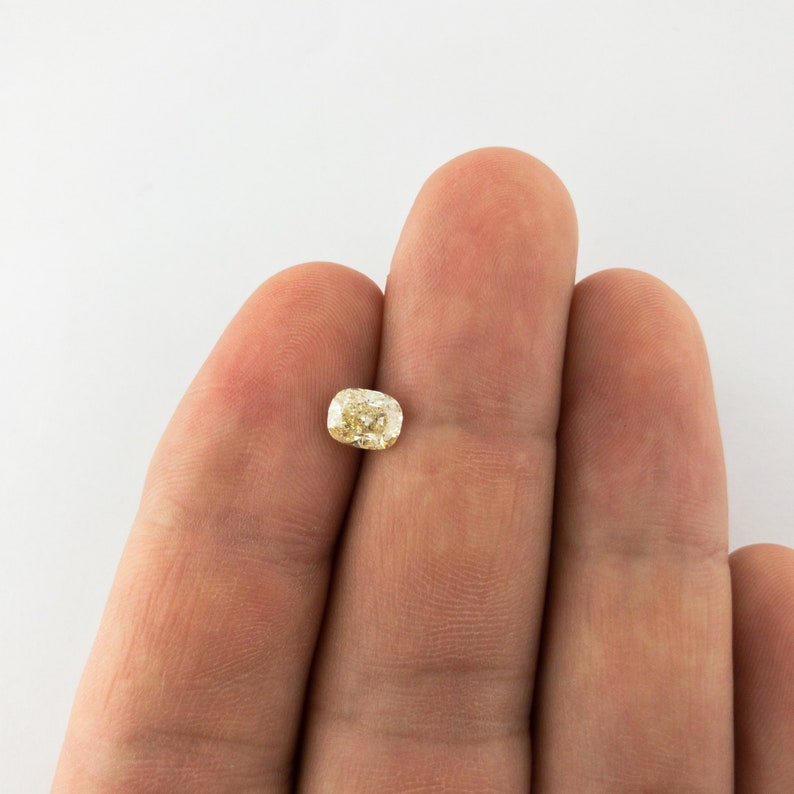 May include: A light yellow cushion-cut gemstone held between two fingers.