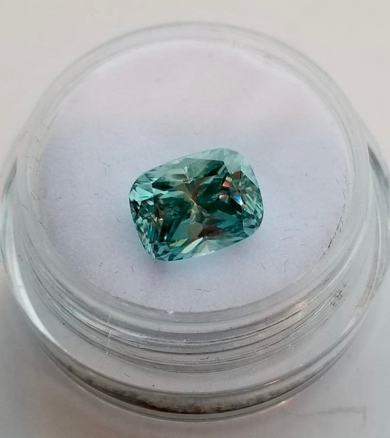 May include: A teal-coloured cushion-cut gemstone with iridescent flashes, set in a clear plastic container with a white interior. The gem's facets reflect light, creating a sparkling effect.