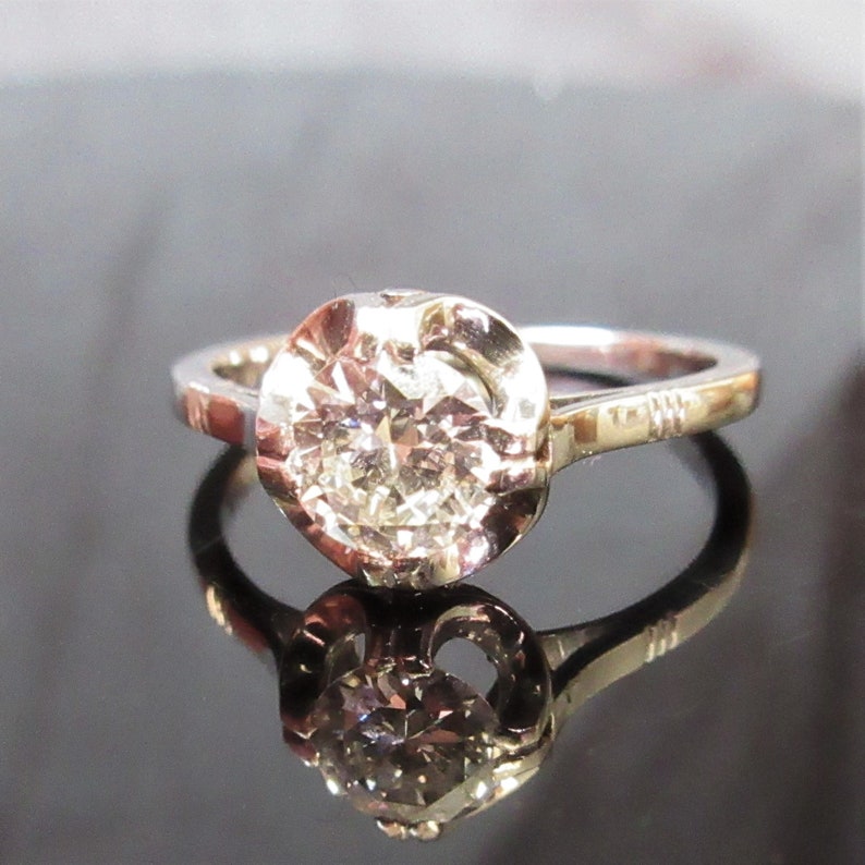 May include: A silver ring with a large, round, sparkling diamond center stone. The diamond is set in a bezel setting, and the band has a simple, elegant design. The ring is displayed on a reflective surface.
