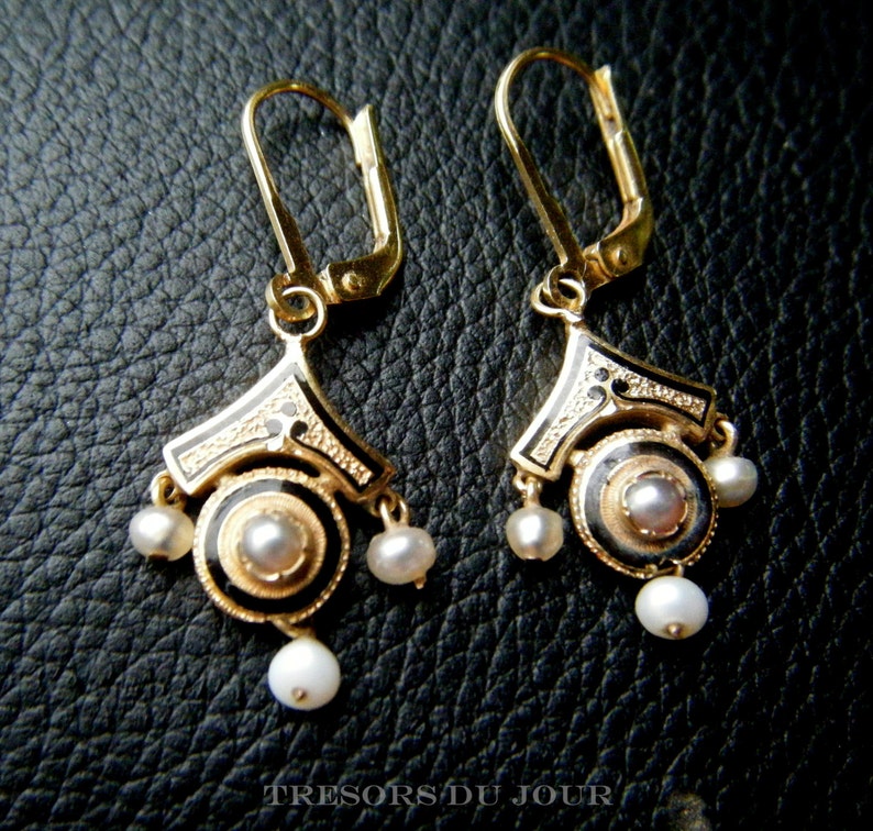 May include: A pair of gold earrings with black enamel and pearl accents. The earrings have a geometric design with a circular center and dangle from a hook.