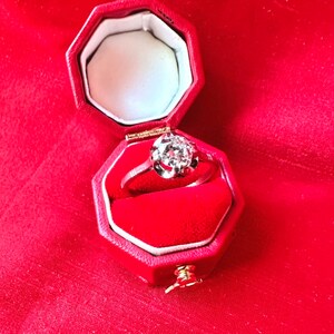 May include: A diamond ring in a red octagonal box. The ring features a large, round-cut diamond set in a silver band. The box is lined with red velvet and sits on a red fabric surface. The ring is a classic piece of jewelry.