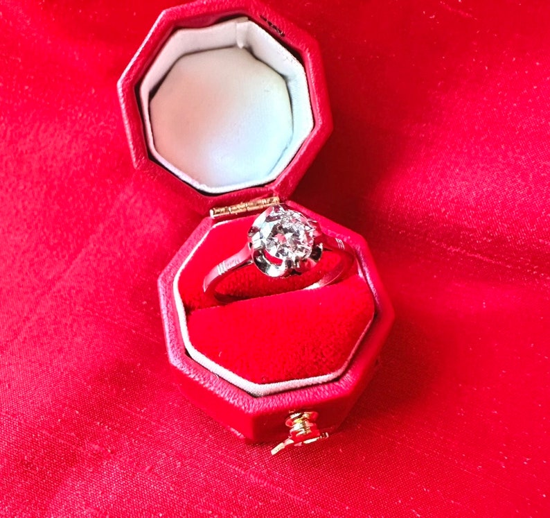 May include: A diamond ring in a red octagonal box. The ring features a large, round-cut diamond set in a silver band. The box is lined with red velvet and sits on a red fabric surface. The ring is a classic piece of jewelry.