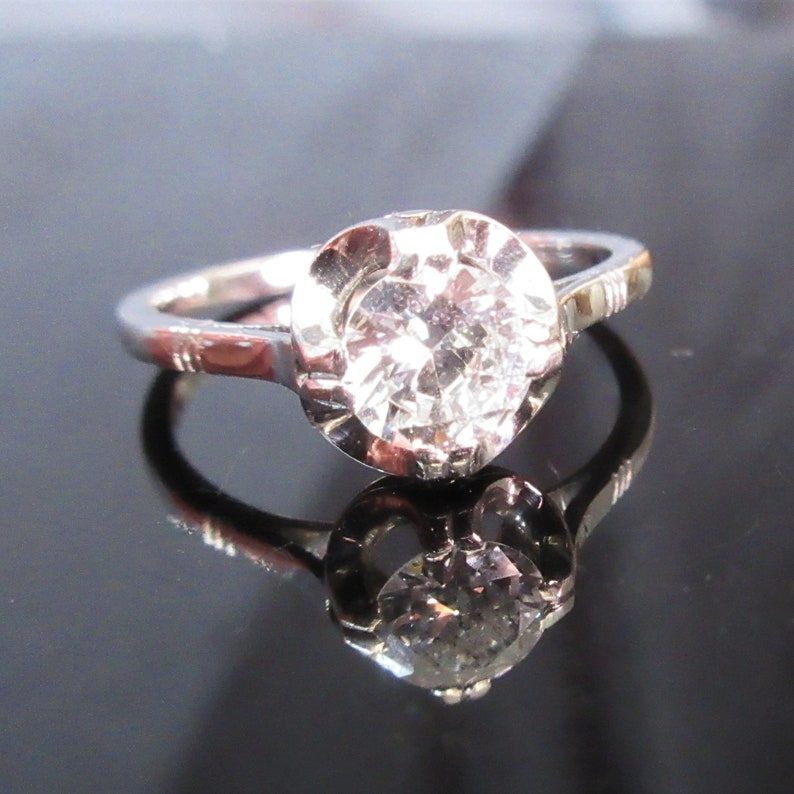 May include: A silver ring with a large, round, clear diamond as the centerpiece. The diamond is set in a bezel setting, and the band has etched details. The ring is displayed on a reflective surface.