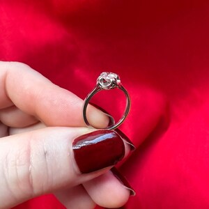 May include: A silver ring with a large, clear gemstone. The ring has a delicate, ornate setting and is held against a red background. The gemstone appears to be round and faceted, catching the light.
