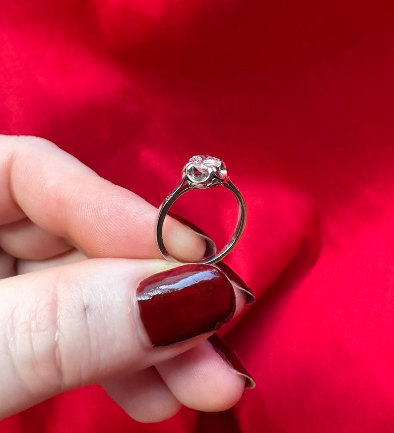 May include: A silver ring with a large, clear gemstone. The ring has a delicate, ornate setting and is held against a red background. The gemstone appears to be round and faceted, catching the light.