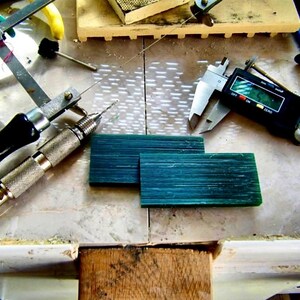 May include: A jeweller's workbench with tools and materials. Includes a digital caliper, two rectangular teal-coloured wax blocks, a hand saw, and a pin vise drill. The tools are arranged on a light-coloured surface.