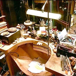 Op de afbeelding: Een rommelige werkbank met gereedschap en benodigdheden voor het maken van sieraden. Er is een lamp, een klok en een verscheidenheid aan gereedschap en materialen op de werkbank.
