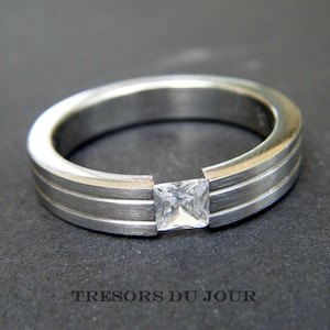 May include: A silver ring with a square-shaped clear gemstone set in the center. The ring has a double band design.