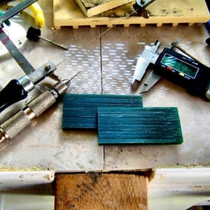 Op de afbeelding: Een close-up van een werkbank met gereedschap voor het maken van sieraden. Er zijn twee groene wasblokken, een boor, een passer, een liniaal en een digitale schuifmaat. De schuifmaat geeft "0.00" aan.