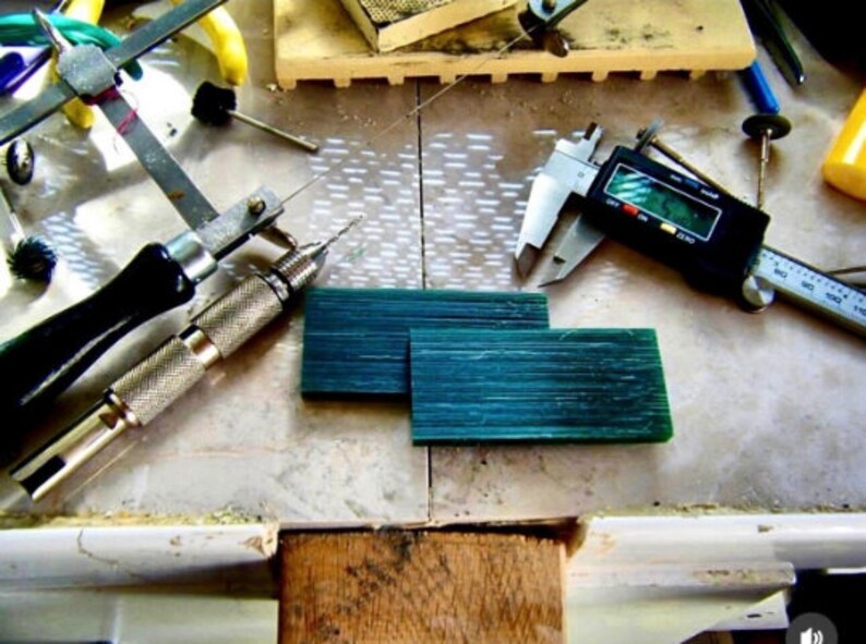 Op de afbeelding: Een close-up van een werkbank met gereedschap voor het maken van sieraden. Er zijn twee groene wasblokken, een boor, een passer, een liniaal en een digitale schuifmaat. De schuifmaat geeft "0.00" aan.