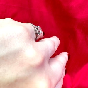 May include: A silver ring with a decorative filigree design and a central clear stone. The ring is set against a vibrant red background, creating a striking contrast. The ring's intricate details are visible.