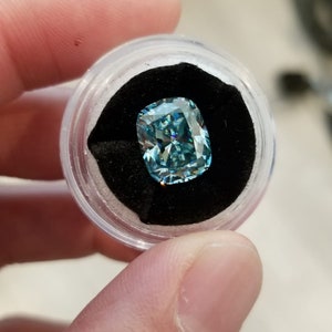 May include: A light blue cushion-cut gemstone, presented in a clear, round container with a black velvet lining. The gemstone's facets catch the light. The container is held in a hand.