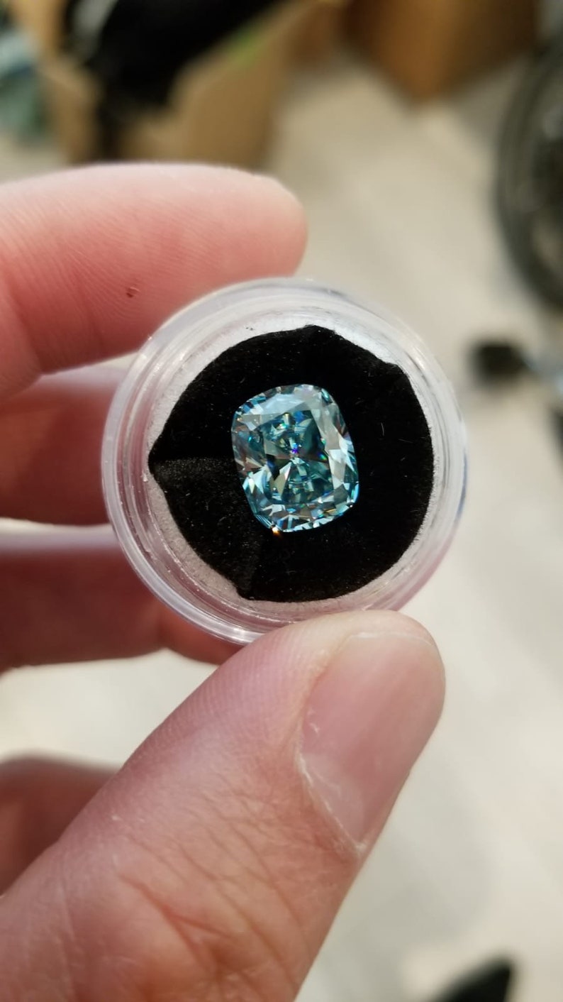 May include: A light blue cushion-cut gemstone, presented in a clear, round container with a black velvet lining. The gemstone's facets catch the light. The container is held in a hand.