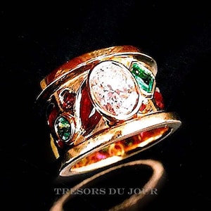 May include: A gold ring with a large, round, clear diamond surrounded by smaller red and green gemstones. The ring is set in a wide band with a textured surface.