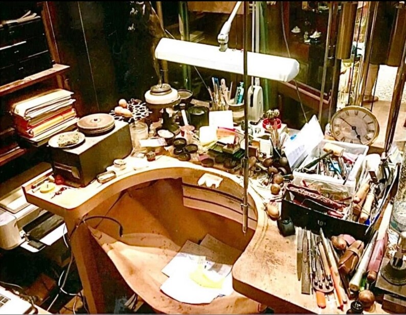 May include: A workbench filled with tools, materials, and a vintage clock. The workspace includes a lamp, stacks of papers, and various small containers. The scene suggests a workshop or studio environment, with a focus on craftsmanship.