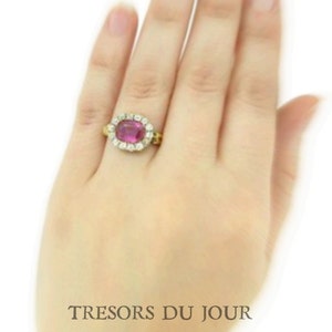 Peut inclure: Une bague en or orn&eacute;e d'une grosse pierre pr&eacute;cieuse ovale de couleur magenta, entour&eacute;e de petits diamants transparents. La bague est port&eacute;e sur une main, avec le texte "TRESORS DU JOUR" en bas.