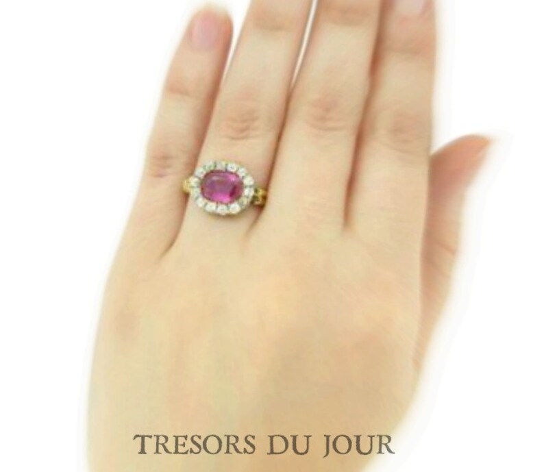Peut inclure: Une bague en or orn&eacute;e d'une grosse pierre pr&eacute;cieuse ovale de couleur magenta, entour&eacute;e de petits diamants transparents. La bague est port&eacute;e sur une main, avec le texte "TRESORS DU JOUR" en bas.