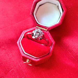 May include: A diamond ring in a red octagonal jewelry box. The ring features a large, round-cut diamond set in a silver band. The box is open, revealing a white interior and red velvet lining. The background is a textured red fabric.