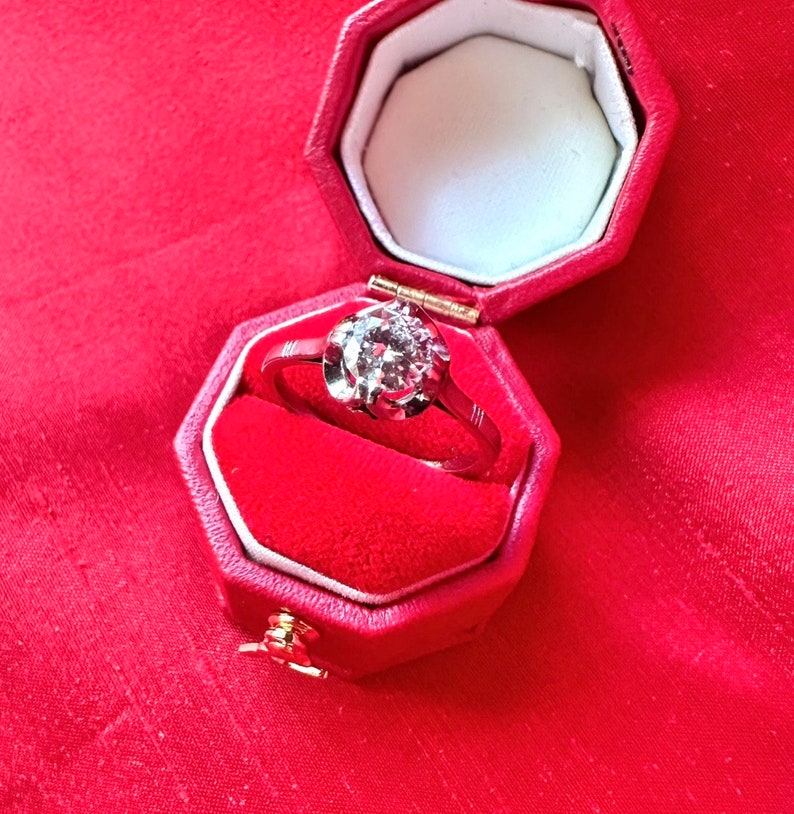 May include: A diamond ring in a red octagonal jewelry box. The ring features a large, round-cut diamond set in a silver band. The box is open, revealing a white interior and red velvet lining. The background is a textured red fabric.