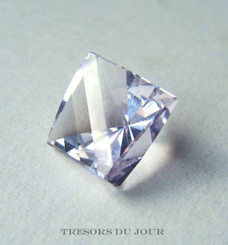 May include: A clear, faceted, square-shaped gemstone with a pale lavender hue. The gemstone is cut in a step-cut style, creating a series of flat facets that reflect light.
