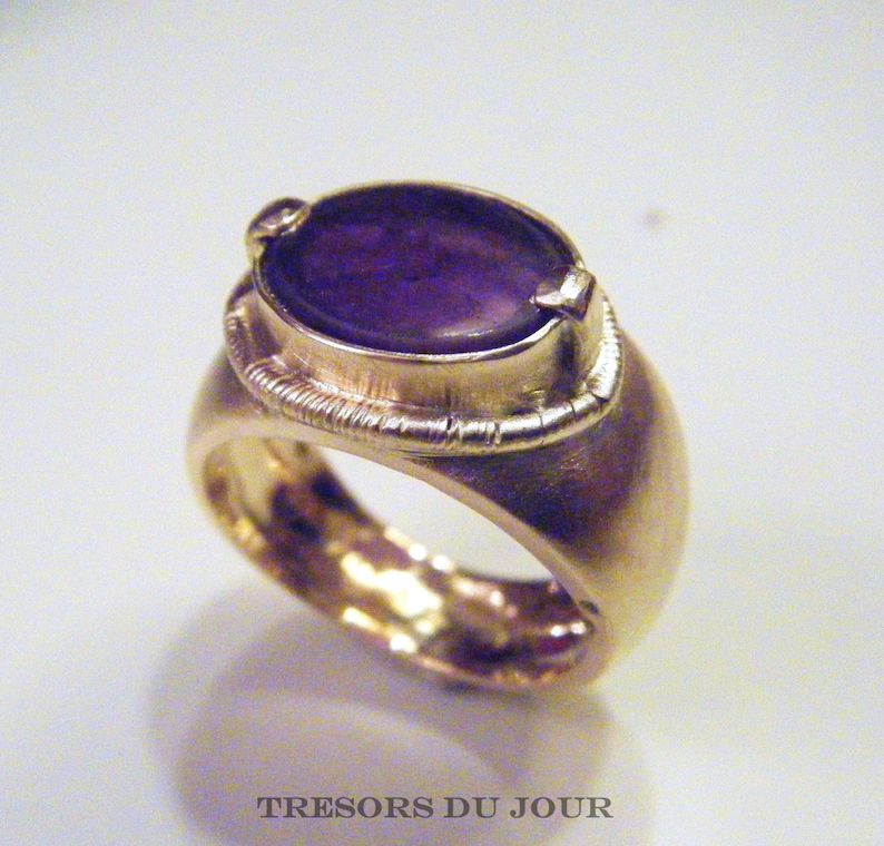 Op de afbeelding: Een gouden ring met een paarse edelsteen in een bezel-setting. De ring heeft een getextureerde band.
