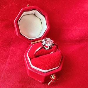 May include: A diamond ring in a red velvet octagonal box. The ring features a large, round-cut diamond set in a silver or white gold band. The box is open, revealing a white interior and a gold clasp. The background is a red fabric.