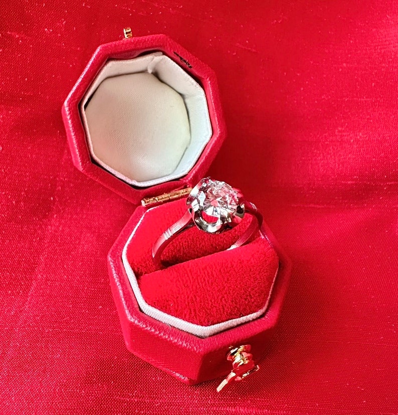 May include: A diamond ring in a red velvet octagonal box. The ring features a large, round-cut diamond set in a silver or white gold band. The box is open, revealing a white interior and a gold clasp. The background is a red fabric.