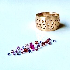 Op de afbeelding: Een gouden ring met een uniek ontwerp met een patroon van kleine cirkels. De ring is omringd door een strooiing van kleurrijke edelstenen, waaronder roze, rode en blauwe.