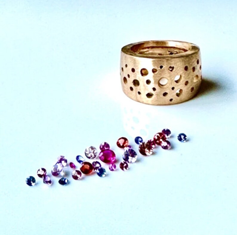 Op de afbeelding: Een gouden ring met een uniek ontwerp met een patroon van kleine cirkels. De ring is omringd door een strooiing van kleurrijke edelstenen, waaronder roze, rode en blauwe.