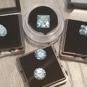 May include: A selection of light blue gemstones in different shapes, including oval, square, and rectangular cuts, presented in individual black cases. Two round gemstones are also visible. The gems are set against a black velvet background.