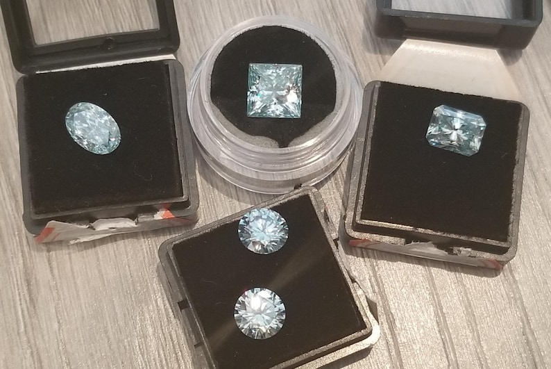 May include: A selection of light blue gemstones in different shapes, including oval, square, and rectangular cuts, presented in individual black cases. Two round gemstones are also visible. The gems are set against a black velvet background.
