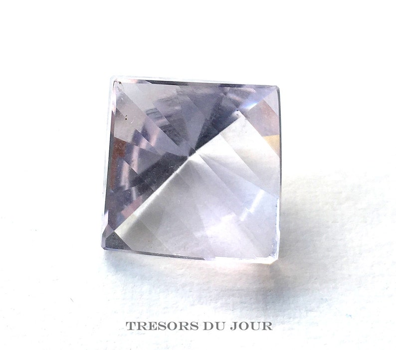 May include: A clear, faceted, square-shaped gemstone with a pale lavender hue. The gemstone is cut into a pyramid shape with a pointed top.