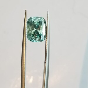 May include: A light teal cushion-cut gemstone is held by tweezers against a white backdrop. The gemstone's facets reflect light, creating a sparkling effect. The tweezers are silver-coloured.