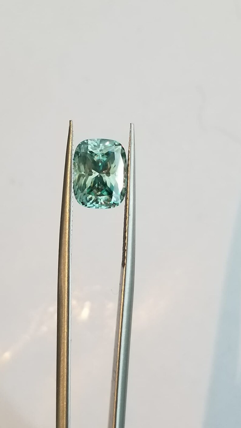 May include: A light teal cushion-cut gemstone is held by tweezers against a white backdrop. The gemstone's facets reflect light, creating a sparkling effect. The tweezers are silver-coloured.