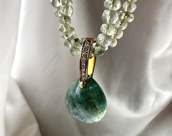 Può includere: Una collana di pietre preziose con un pendente a forma di goccia in una montatura d'oro, impreziosita da piccoli diamanti. Il pendente mostra tonalità verdi e blu. La collana ha due fili di perline sfaccettate verde chiaro.