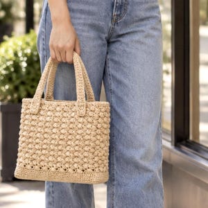 May include: A beige, rectangular, hand-crocheted tote bag featuring a textured, repeating popcorn stitch pattern throughout the body, a flat base, and two matching crocheted top handles for carrying by hand.