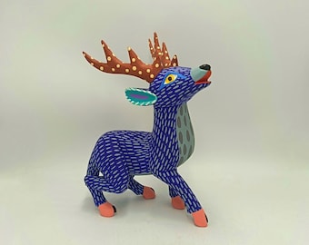 May include: A vibrantly painted wooden deer figurine. The deer features a blue body with white linear patterns, brown antlers with yellow spots, and pink hooves. Its head is raised, and it has a gray patch on its side. A colorful decorative item.
